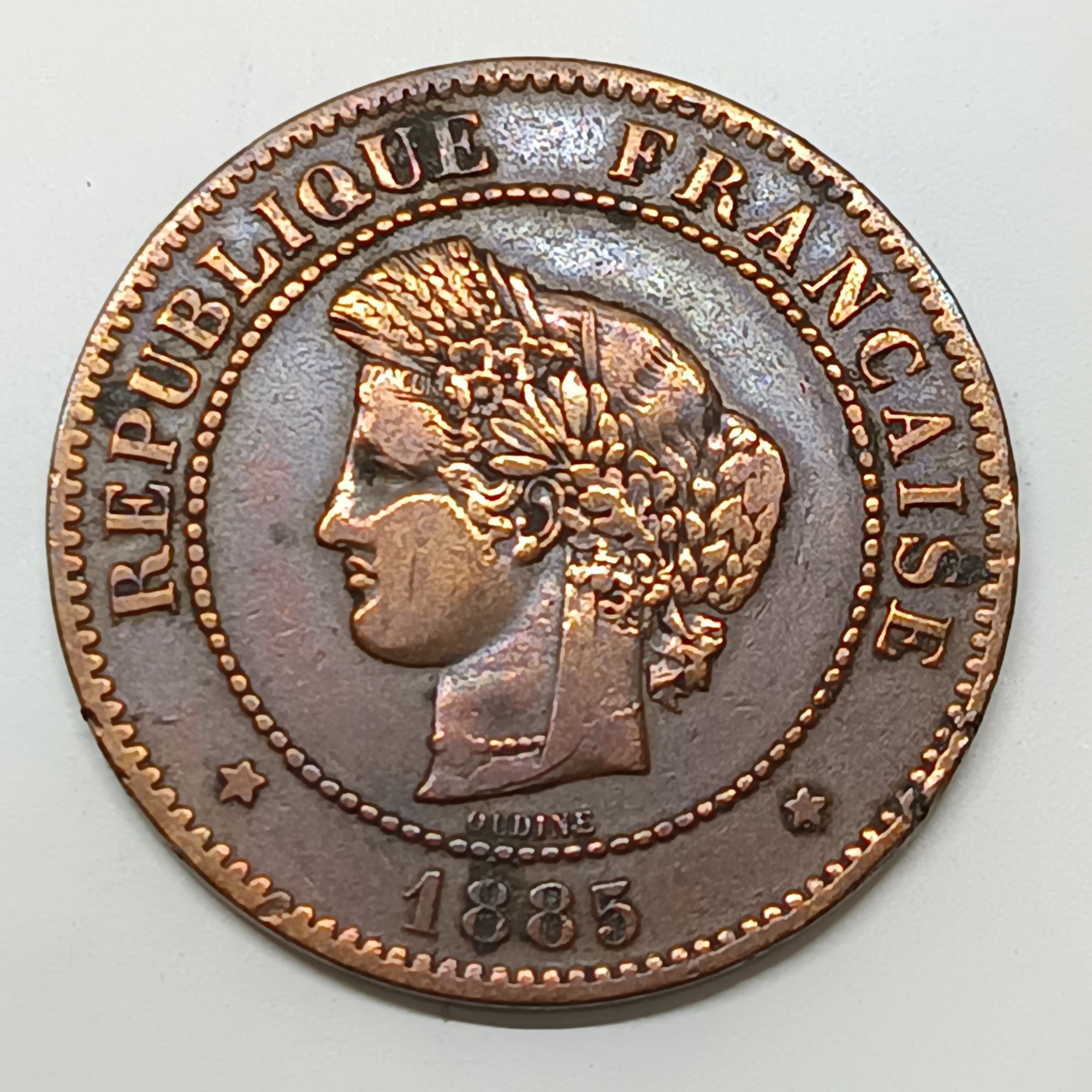 Revers ​Le revers présente au centre la valeur faciale "5 CENTIMES" sur deux lignes, surmontant l'année de frappe "1885". Juste en dessous de la date se trouve la lettre d'atelier "A", indiquant que la pièce a été frappée à la Monnaie de Paris. L'ensemble est entouré d'une couronne composée d'une branche de chêne à gauche et d'une branche d'olivier à droite, symbolisant respectivement la force et la paix. La devise de la République française, "LIBERTÉ • ÉGALITÉ • FRATERNITÉ", est inscrite en cercle le long du listel.