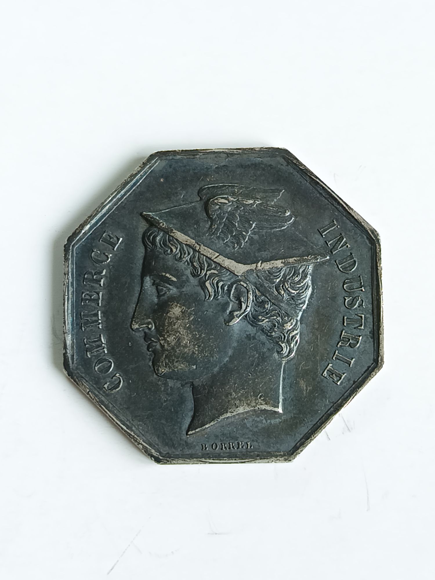 Ce jeton est un objet numismatique français émis en 1854 pour la Caisse d'escompte de Troyes. Voici les détails que j'ai pu rassembler à son sujet : Description du jeton : * Type : Jeton octogonal * Métal : Argent (AR) * Poids : Généralement autour de 12,77 grammes. * Diamètre : Environ 30,5 mm. * Tranche : Lisse, avec un poinçon en forme de main, qui est une marque d'argent. * Avers : On y voit une tête de Mercure tournée vers la gauche, avec la signature "BORREL" en dessous. La titulature est "COMMERCE - INDUSTRIE". * Revers : La titulature est "DEPARTEMENT DE L'AUBE * CAISSE D'ESCOMPTE". Au centre, il y a l'inscription "TROYES" et le millésime "1854". Contexte historique : Le jeton a été émis à une époque de forte croissance économique en France, durant la Révolution industrielle. De nouvelles banques provinciales, souvent appelées "caisses d'escompte", ont été créées pour répondre aux besoins de financement des entreprises et du commerce. Ces établissements jouaient un rôle essentiel en facilitant les transactions et en accordant des crédits à court terme. La Caisse d'escompte de Troyes était l'une de ces banques. Valeur et rareté : La valeur d'un jeton dépend de son état de conservation (patine, rayures, chocs sur la tranche, etc.). D'après les informations disponibles, un jeton de ce type en bon état de conservation (TTB) a été vendu pour environ 95 € en 2012. Il est important de noter que ce prix est une indication et que la valeur peut varier considérablement en fonction du marché numismatique actuel.